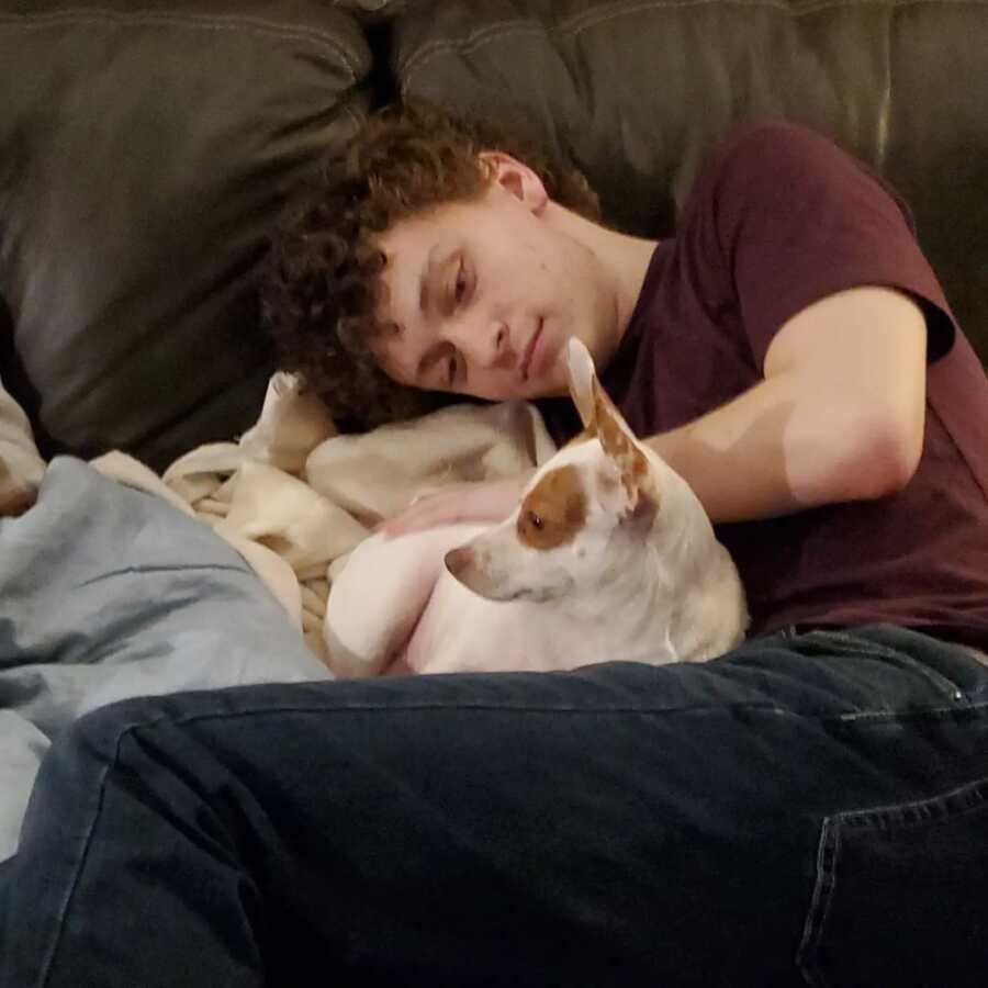 Teen boy lays on couch with little dog curled up against him.