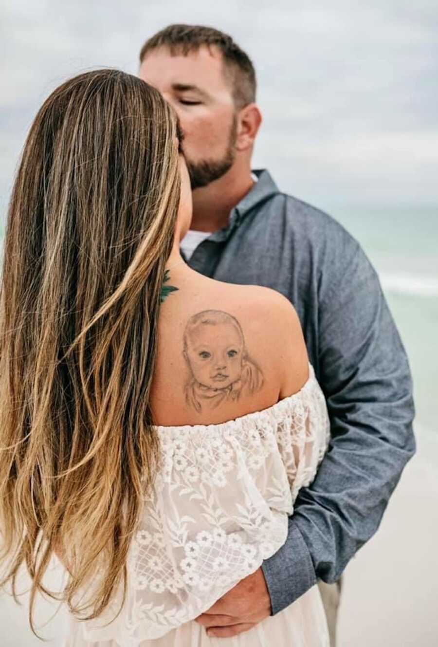 husband holds his wife and kisses her forehead, you can see wife's tattoo of son they lost