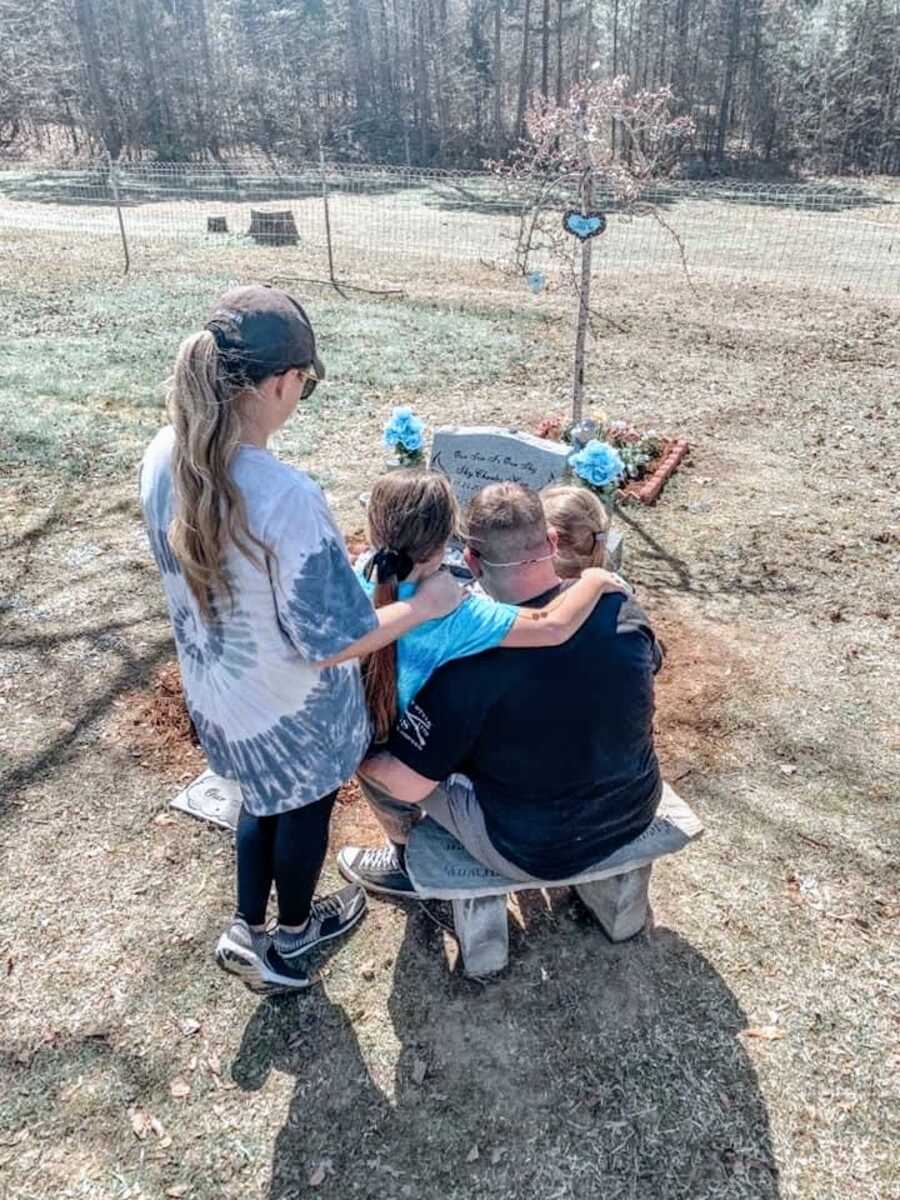 mother, father, and two daughters visit the grave of their son/brother, all of them are holding one another