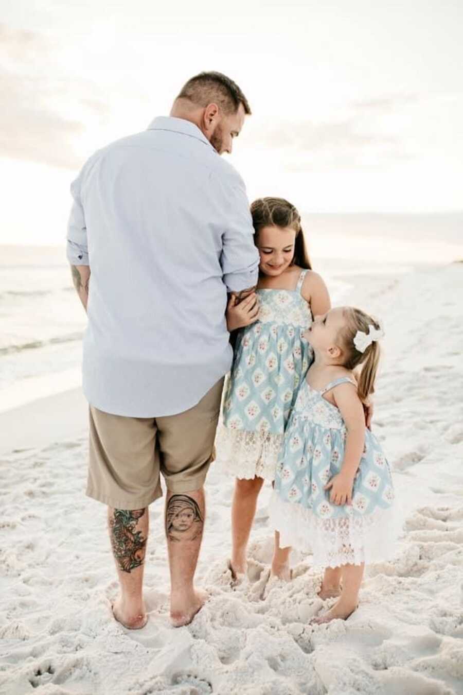dad stands with his two daughters and shows off tattoo of his son who has passed on the back of his leg