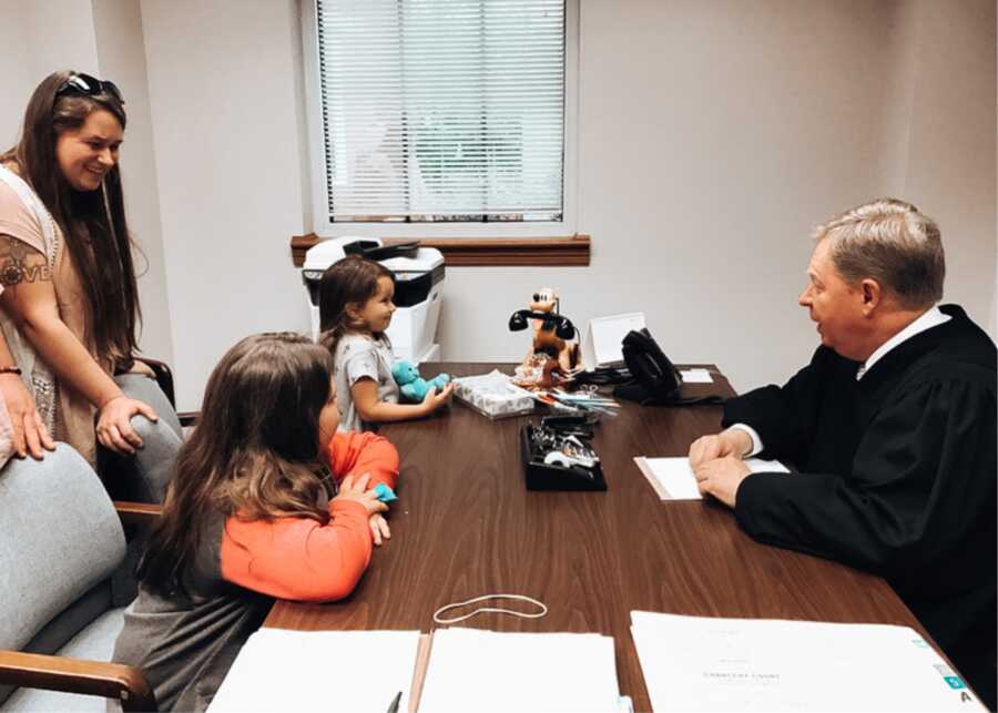 two young girls sit at the judge's desk, mom standing behind them, on adoption day