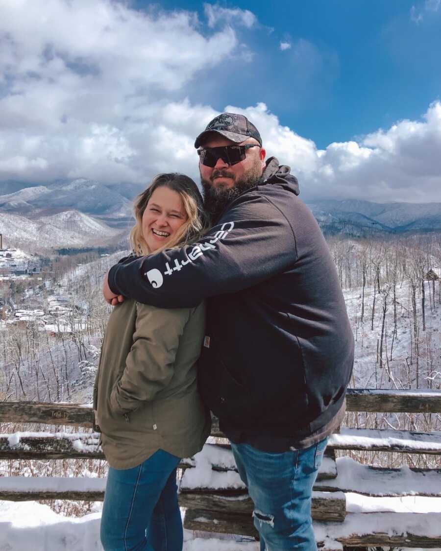 Husband has arms around wife as they stand sideways, taking a picture in front of a snowy mountain landscape. 