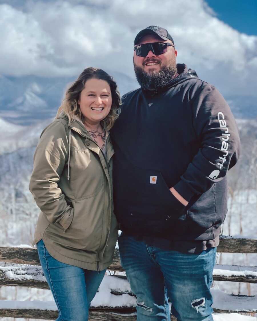 Husband and wife stand side by side with arms around each other in front of a snowy mountain landscape.