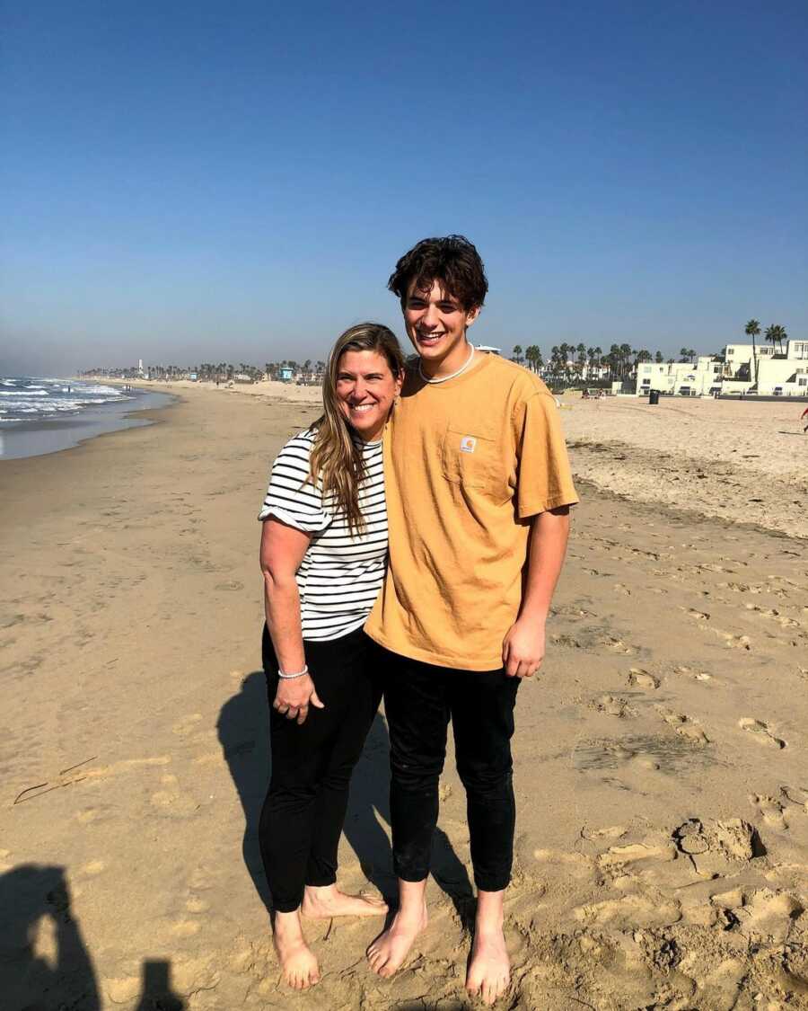 Teen boy and his mom stand with their arms around each other for a picture at the beach.