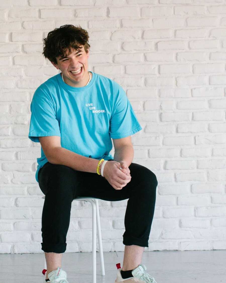 Teen boy wearing blue shirt and black pants sits on a stool in front of a white brick wall, smiling and clasping his hands with his arms resting on his legs. 