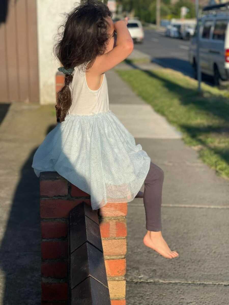 little girl sitting on a brick wall looking out on to the street