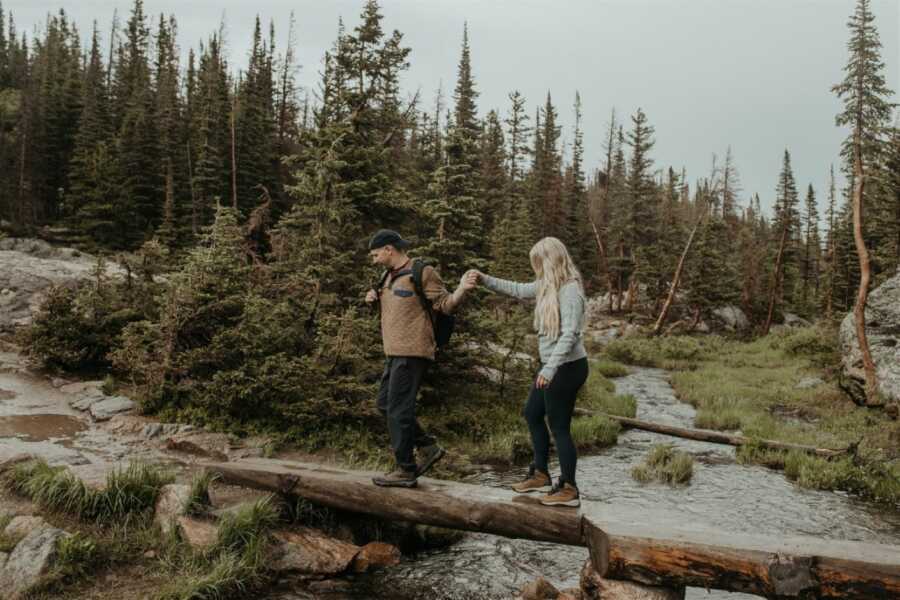 Newlyweds hold hands while walking on a log over a stream during a hike in the forest