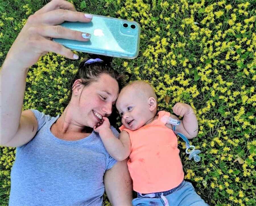 Mom laying on the grass laughing with her baby girl while taking a picture