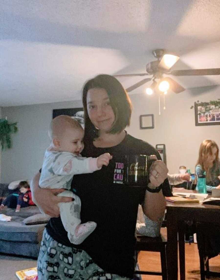 Mom takes a sleepy selfie while holding her daughter in one hand and her coffee mug in another