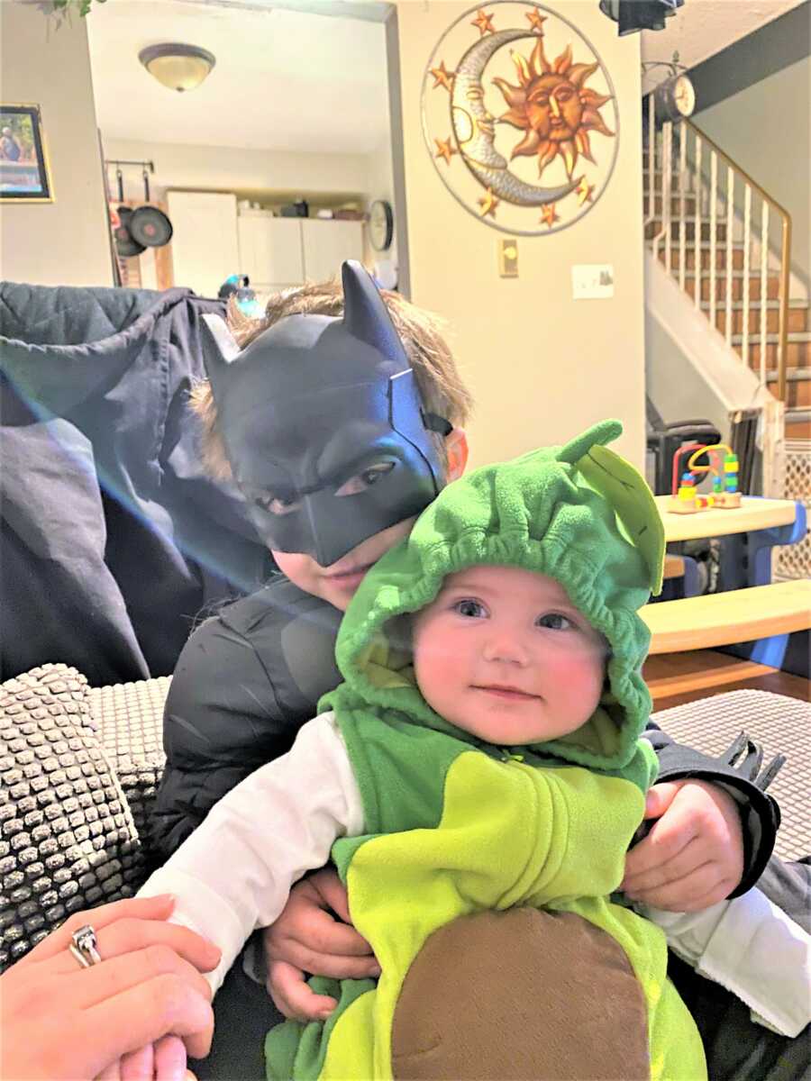 toddle brother dressed as Batman holding his baby sibling dressed as an avocado