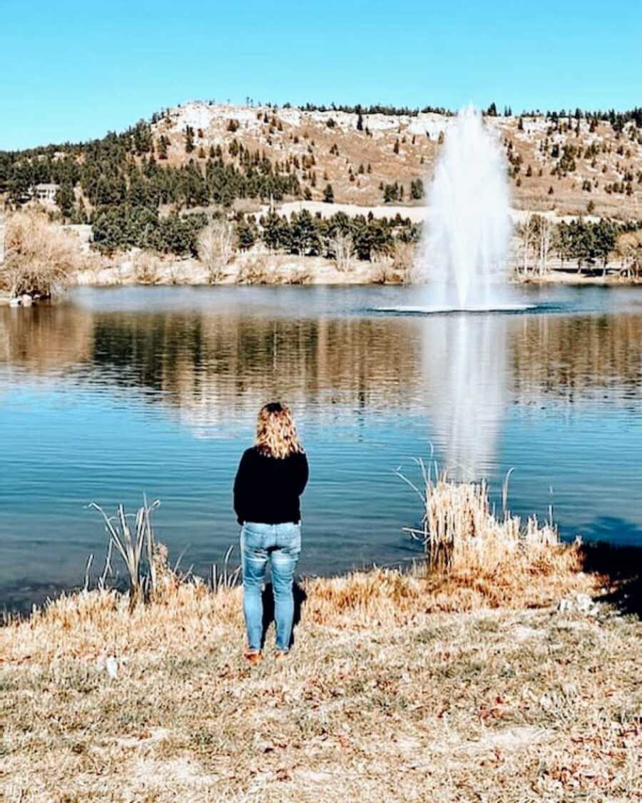 Widow mournfully stares out over a lake while wearing a black shirt and blue jeans
