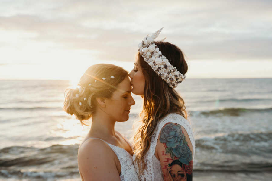 Wedding photography of lesbian couple at the beach kissing the forehead of her bride 