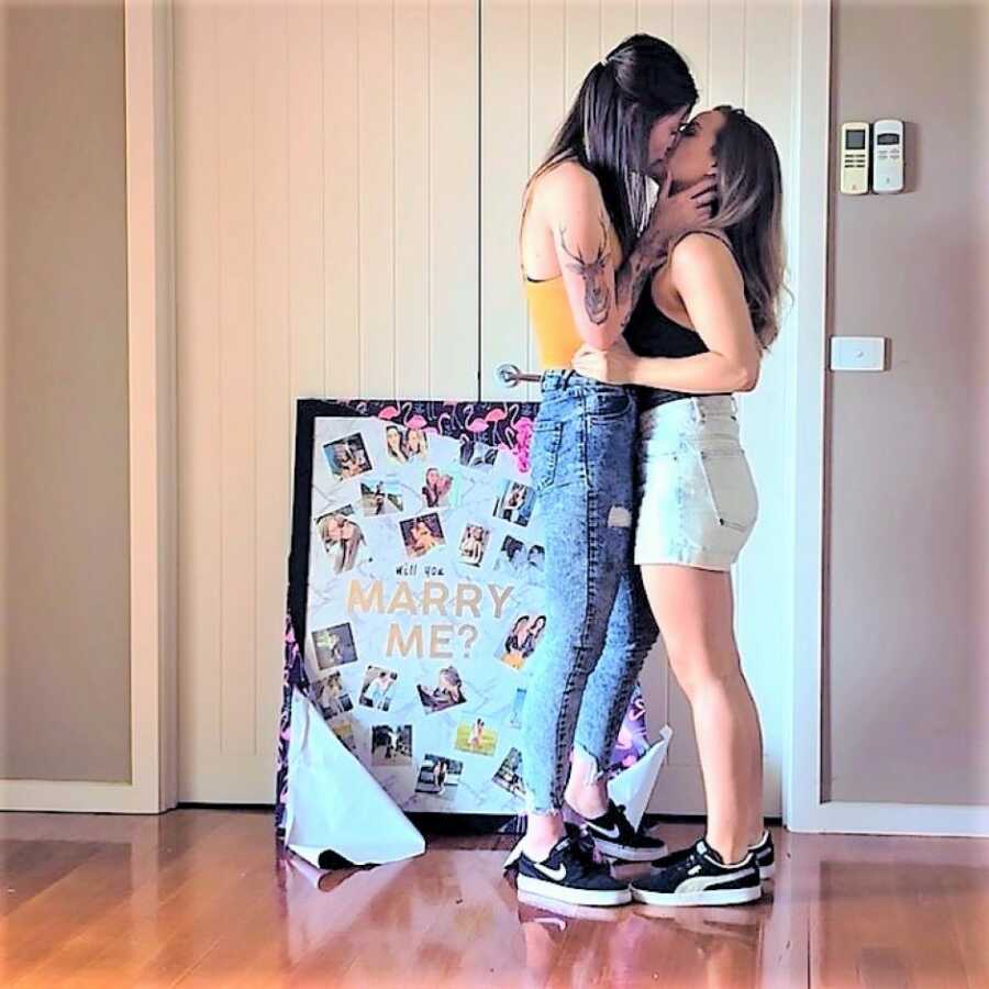 Lesbian couple kissing after marriage proposal with a sign that says "Marry me"
