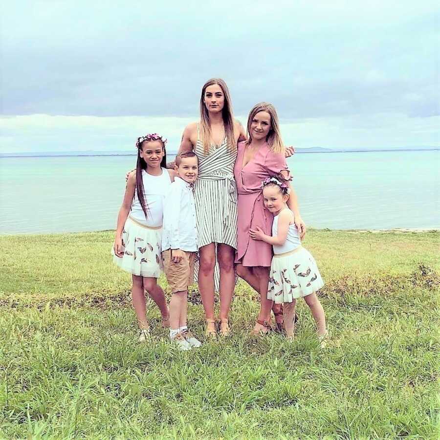 Mom standing next to her three kids and her girlfriend wearing semi-formal outfits with the ocean in the background 