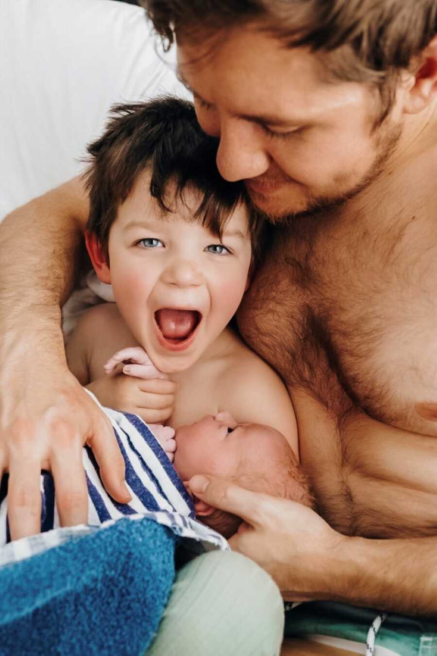 Dad holding his son and newborn son closely while older brother looks thrilled 