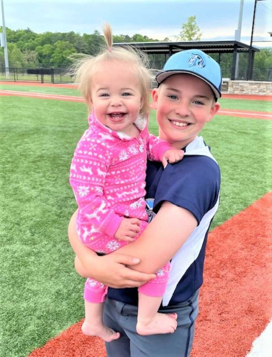 big brother carrying his baby sister at a baseball field 