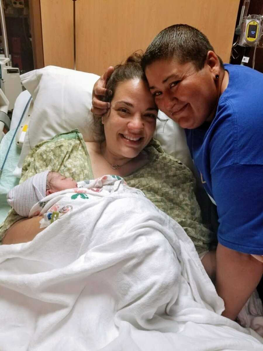 Two moms smile while holding their newborn daughter in the hospital