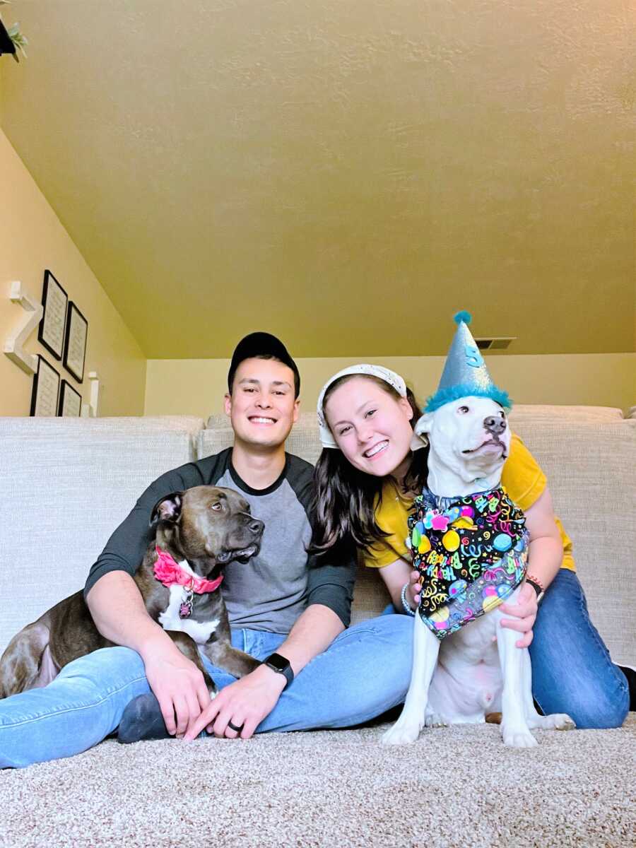 husband and wife sitting on a carpeted floor with a brown dog and a white dog dressed in a birthday costume 
