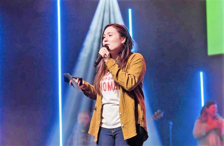 woman holding a mic on a stage with bright lights and a music band in the back