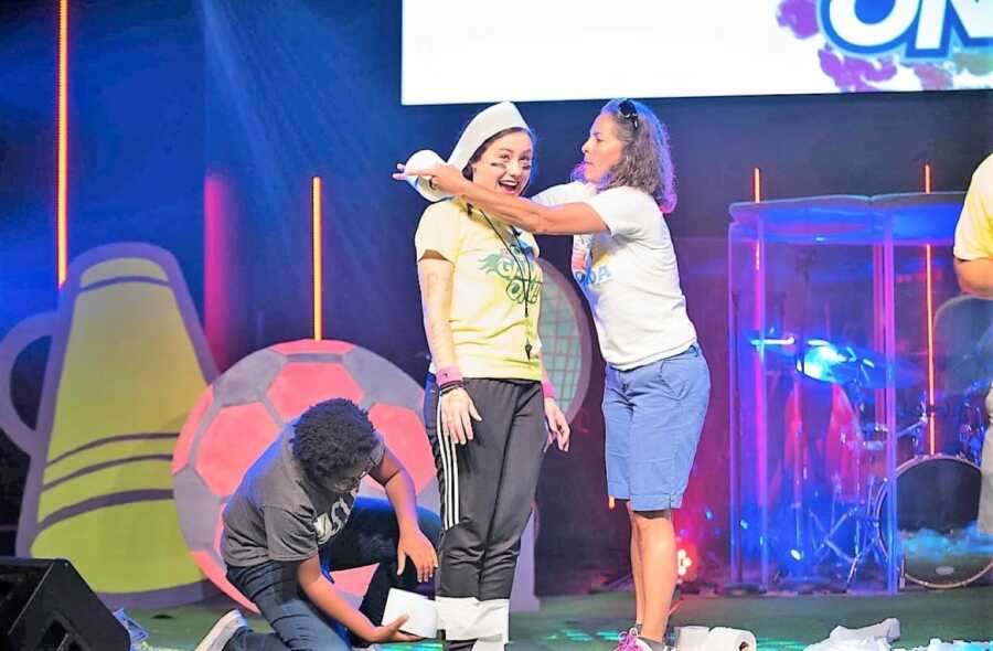 girl looking excited standing on a stage with bright colors and lights while two people wrap her with toilet paper 