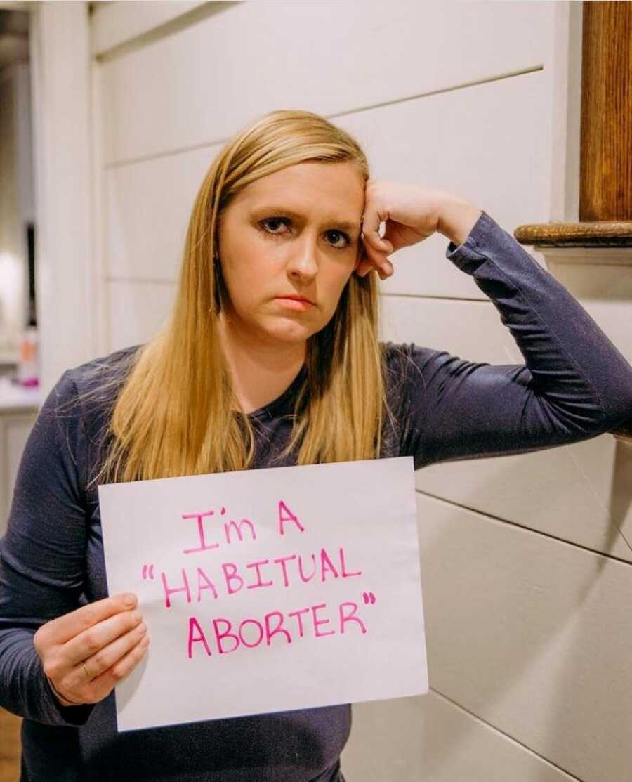 Blonde woman looking serious holds a sign that reads "I'm a 'habitual aborter'"