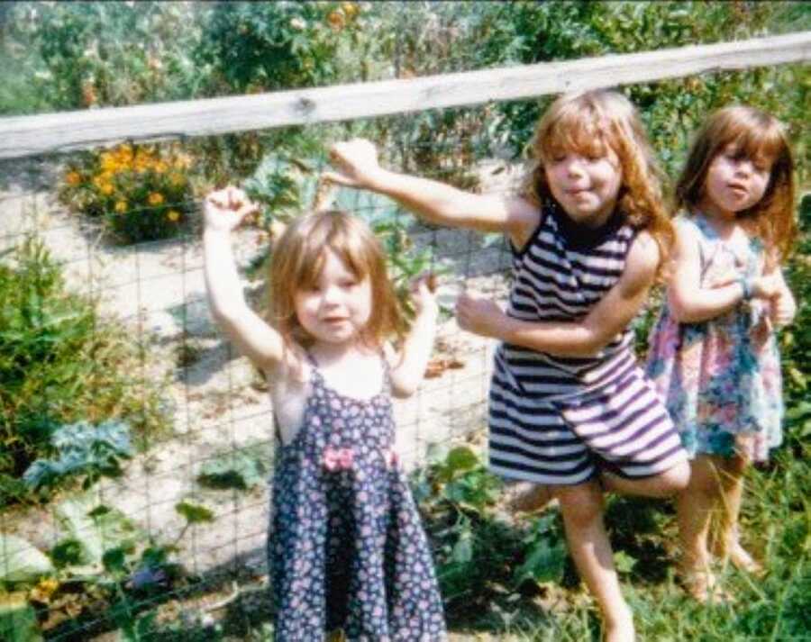 Three sisters in dresses pose for a photo against a fence in their backyard with a garden behind them