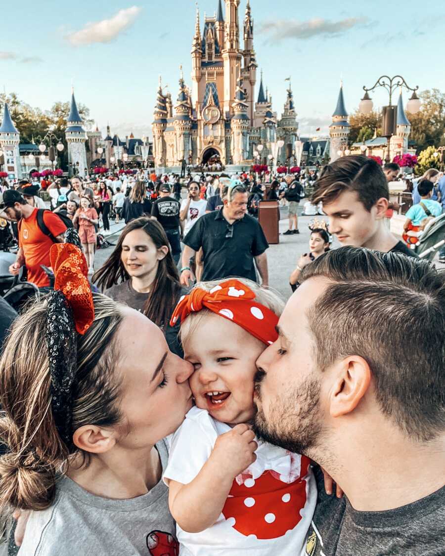 mom and dad at disneyland with their daughter kissing her