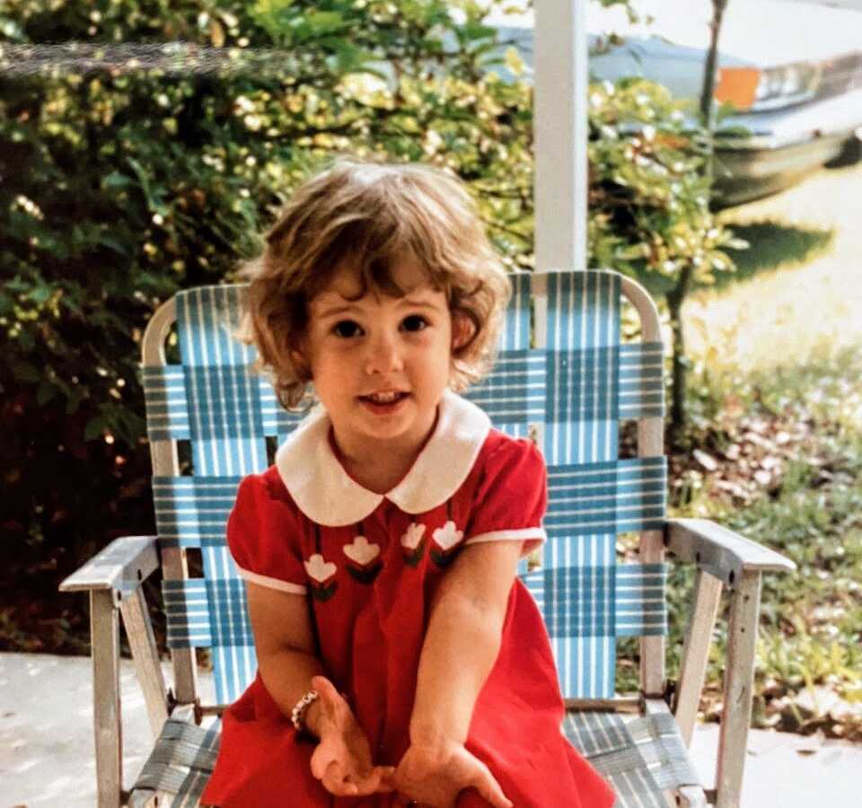Young girl sits on blue outdoor chair while wearing a red dress