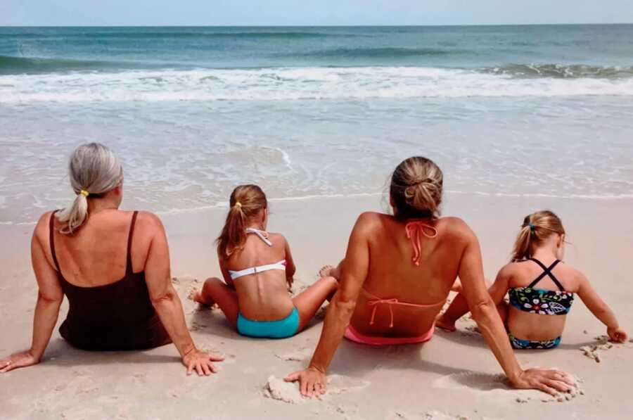 Three generations of women sit in the sand and look out over the water at the beach
