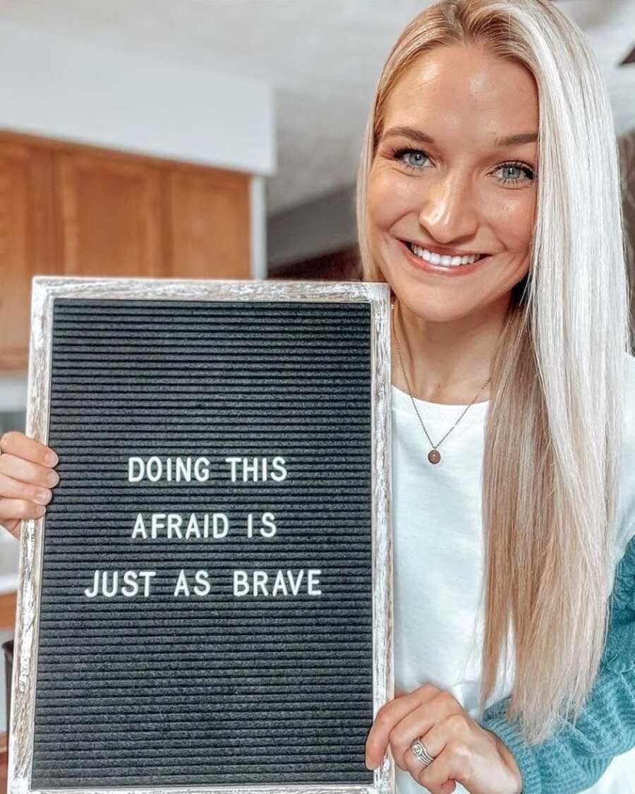 Woman battling infertility smiles for a photo while holding a sign that reads "Doing this afraid is just as brave"