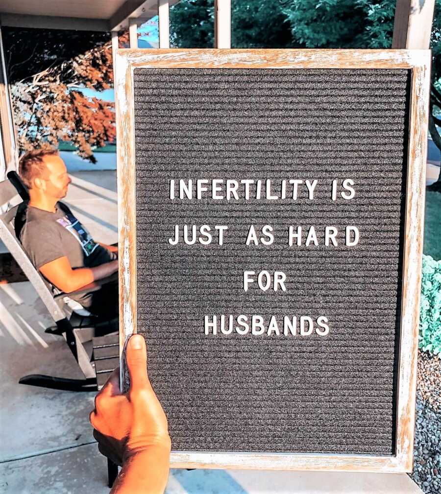 wife holding a sign that says "INFERTILITY IS JUST AS HARD FOR HUSBANDS" with a husband sitting in the background 
