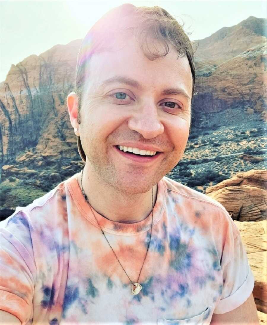 selfie of man smiling on a hike with mountains in the background and the sun 