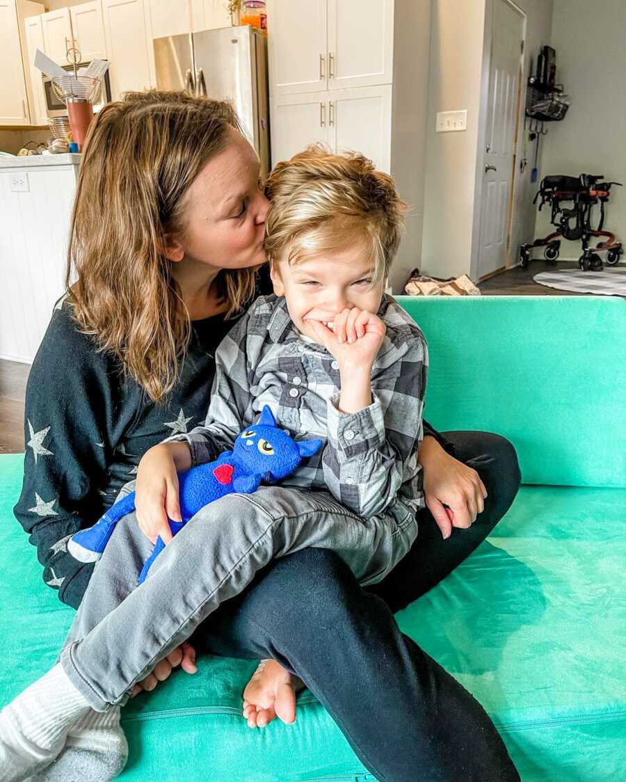Mom kisses her special needs son on the temple while he sits in her lap on the couch