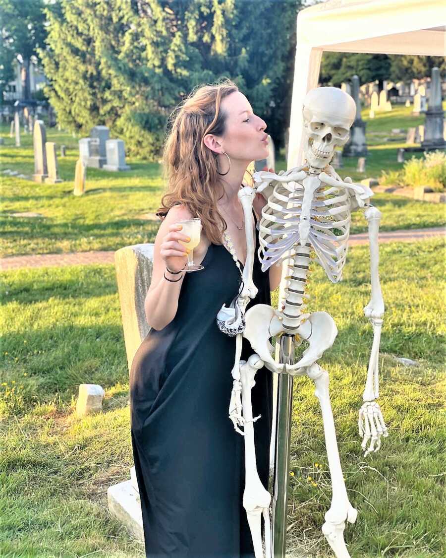 Woman wearing a long black dress and having drink while kissing a fake human skeleton at a cemetery 
