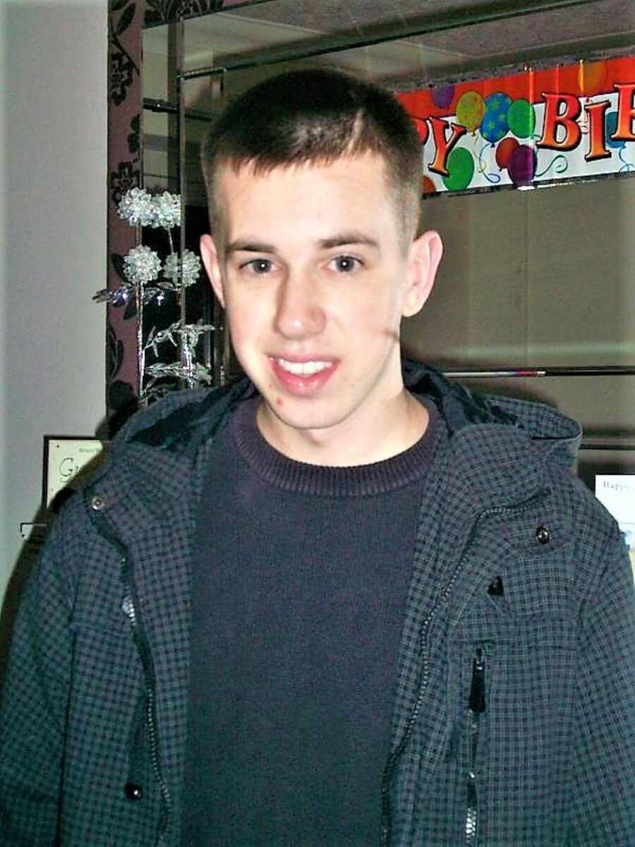 young man with a scar on his head smiling with a "Happy Birthday" sign in the background