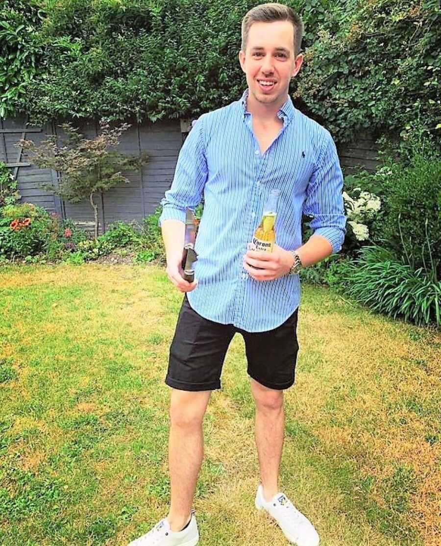 Young man wearing a blue long sleeve shirt and shorts holding a Corona beer