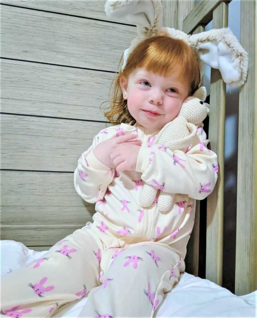 Toddler girl wearing a onesie and bunny ears hugging a stuffed crocheted bunny 