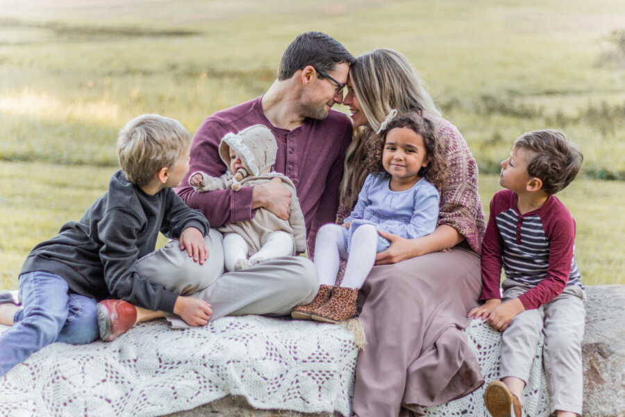 Outdoor family portrait of mom and dad looking at each other while sitting next to their 4 kids