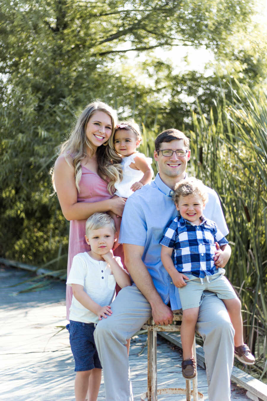 Outdoor family portrait of mom carrying her daughter, dad sitting on a stool with his son on his lap and and their oldest son standing next to them