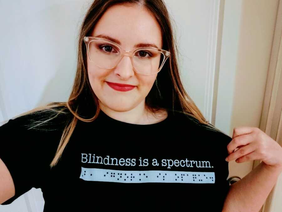 A visually impaired woman wears a black shirt reading "Blindness is a spectrum" in letters and braille