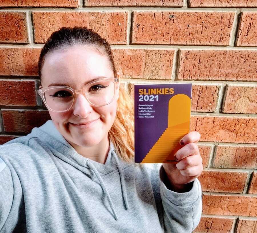 A young woman in a grey sweatshirt and glasses holds up a book in front of a brick wall