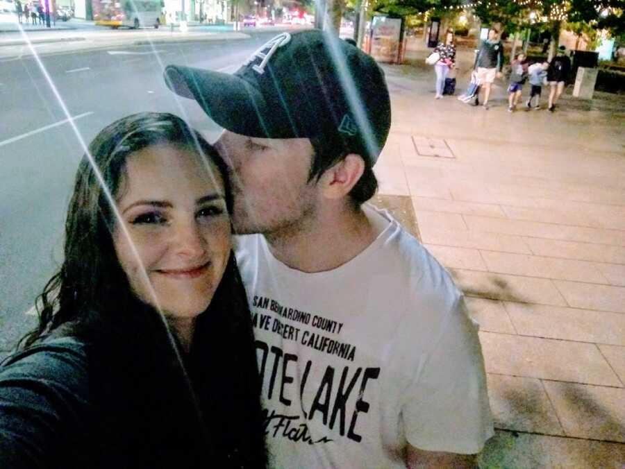 A man wearing a baseball cap kisses his partner on the side of the head