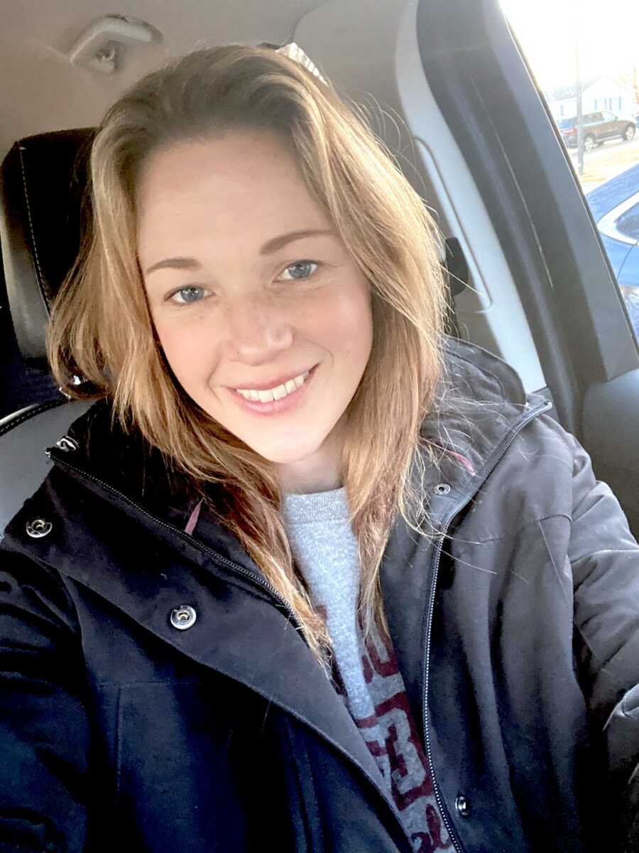 Young woman wearing a grey coat and blue shirt takes a selfie sitting in her car.