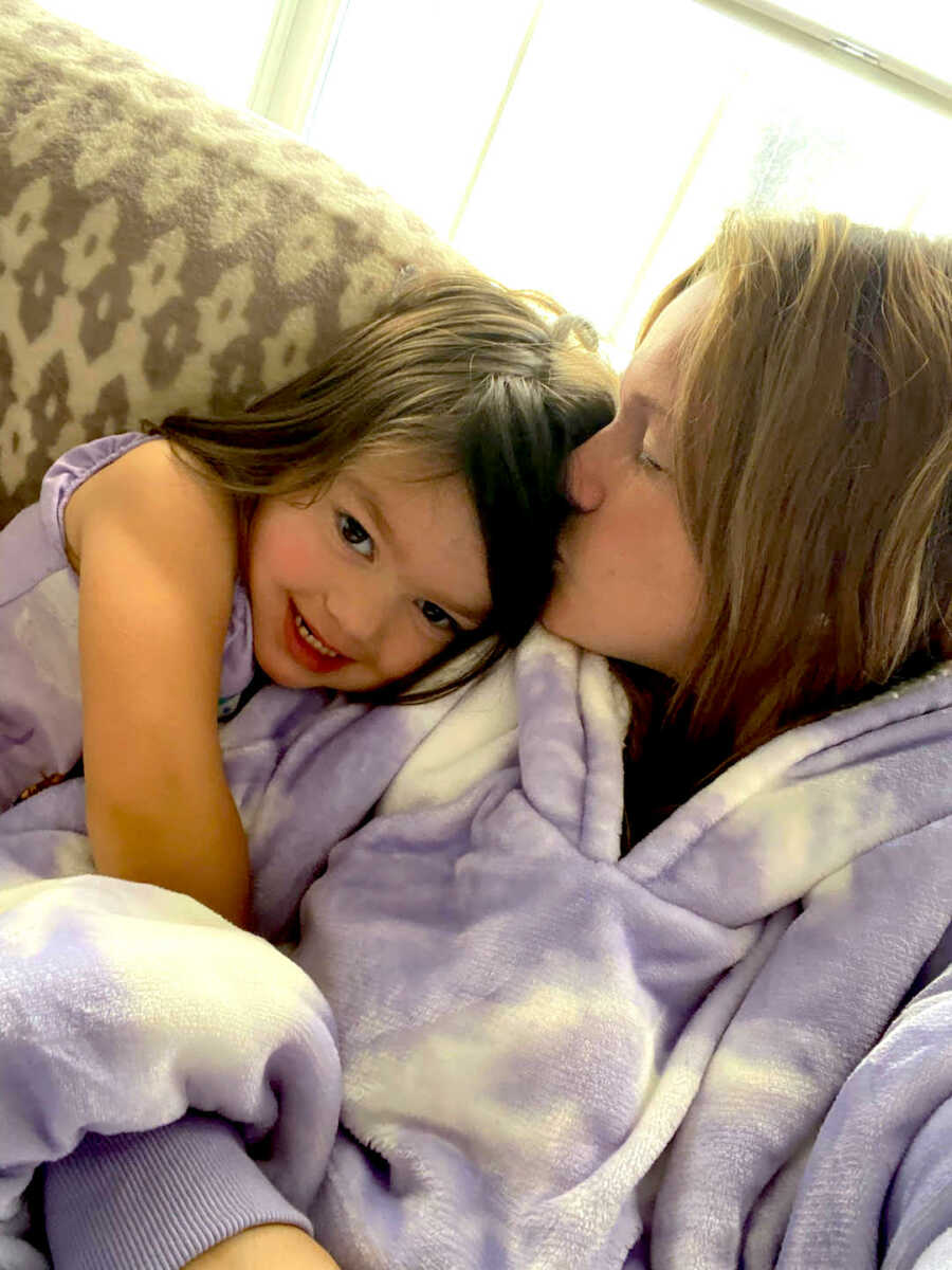 Daughter wraps arms around her mother's arm and rests her head on her shoulder while she kisses her hair.