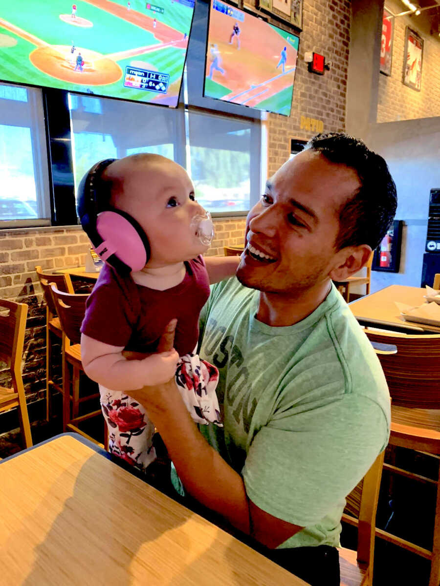 Man in green shirt holds up baby wearing ear muffs.