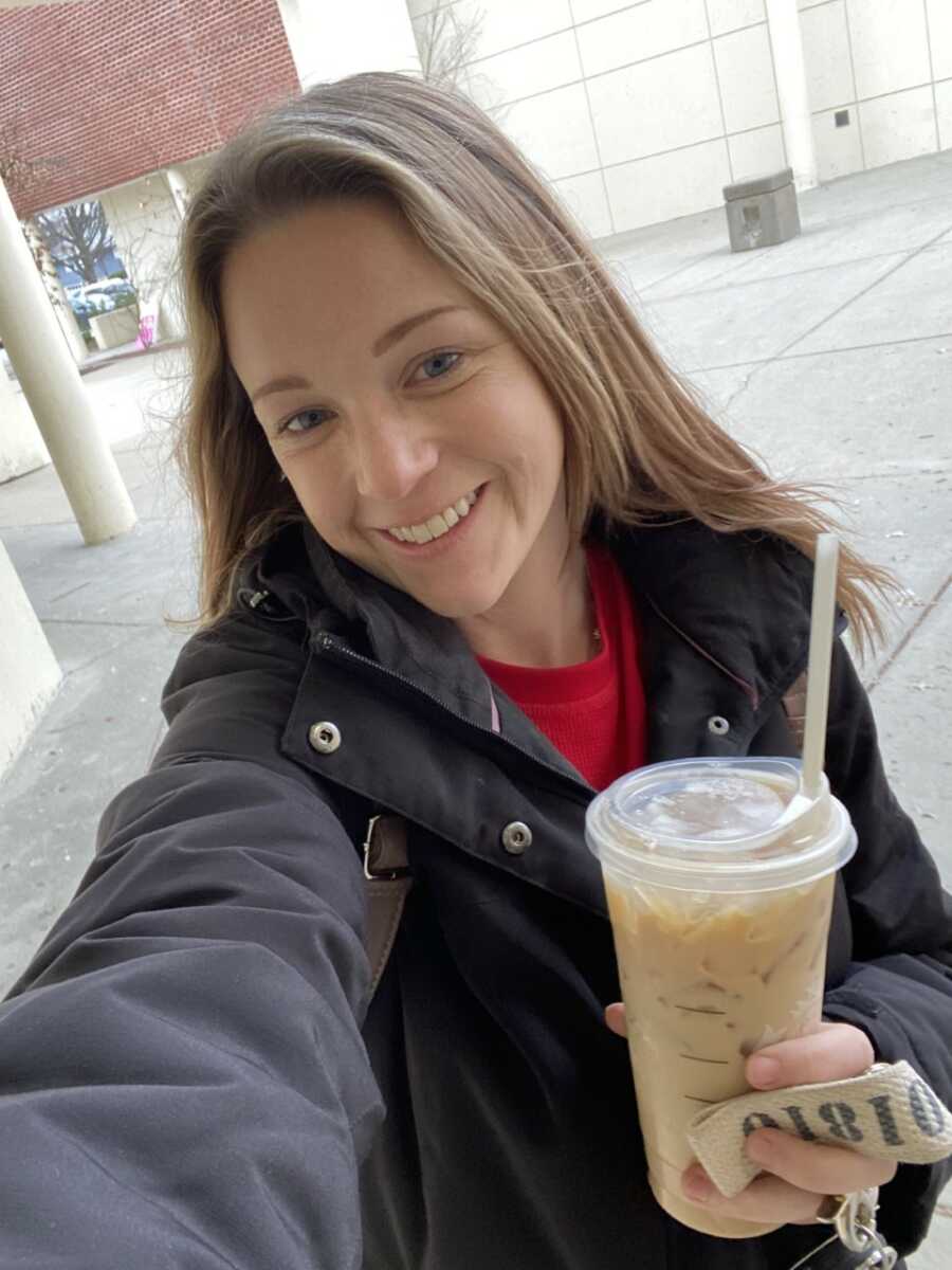 Woman wearing grey coat and red shirt holds up iced coffee and smiles for selfie. 
