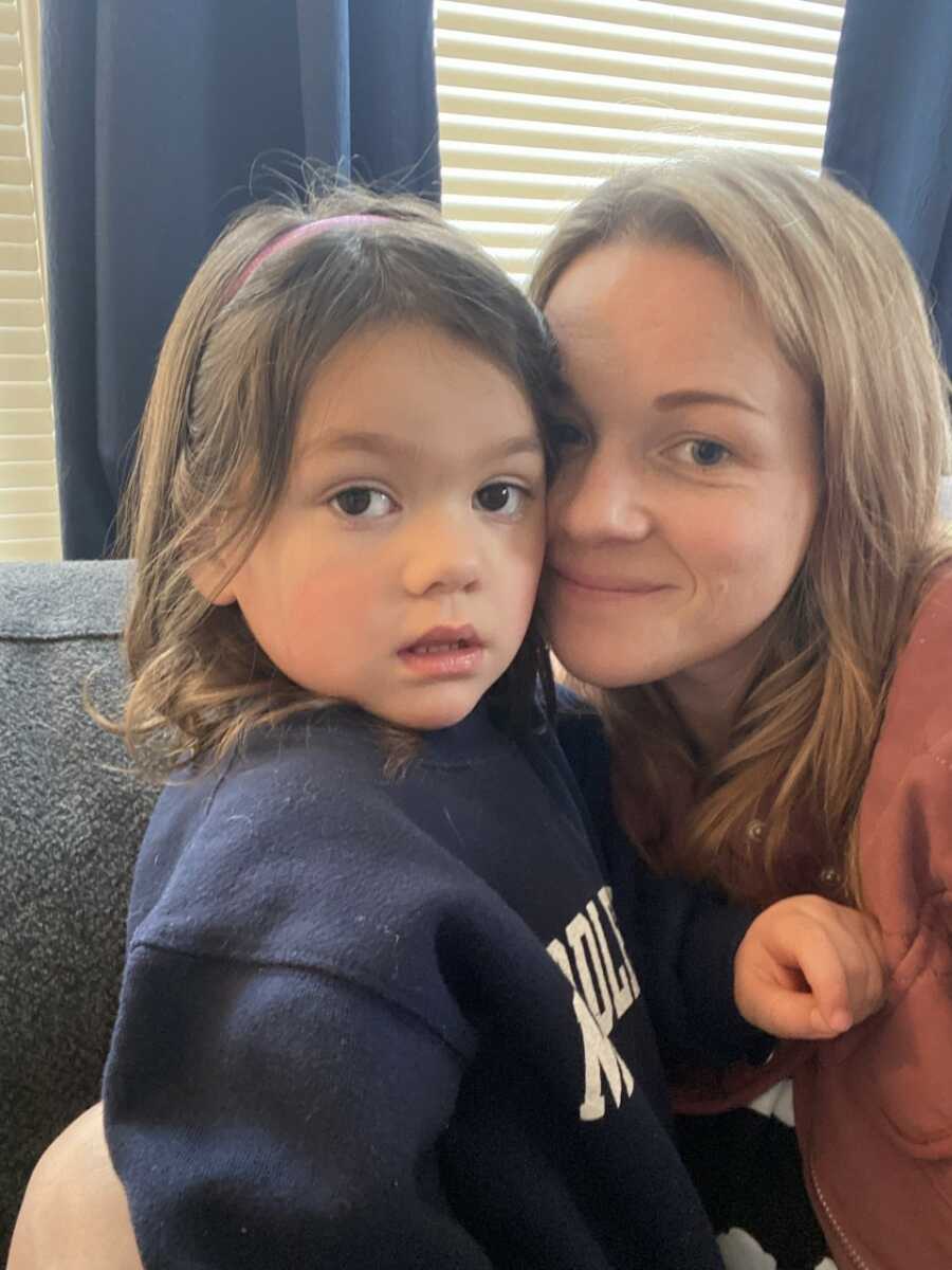 Mom presses face against young daughter's for selfie on the couch together. 