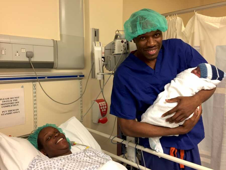 Man proudly holds his newborn child while his wife lays in a hospital bed