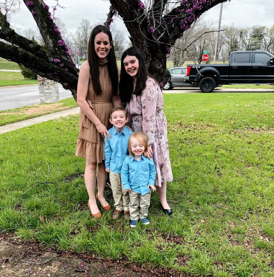 A mom stands with her two young kids outside by a tree