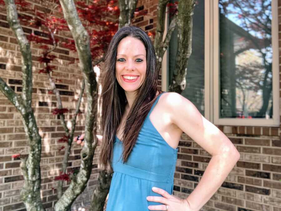 A mom stands by a brick wall and a tree wearing a blue dress